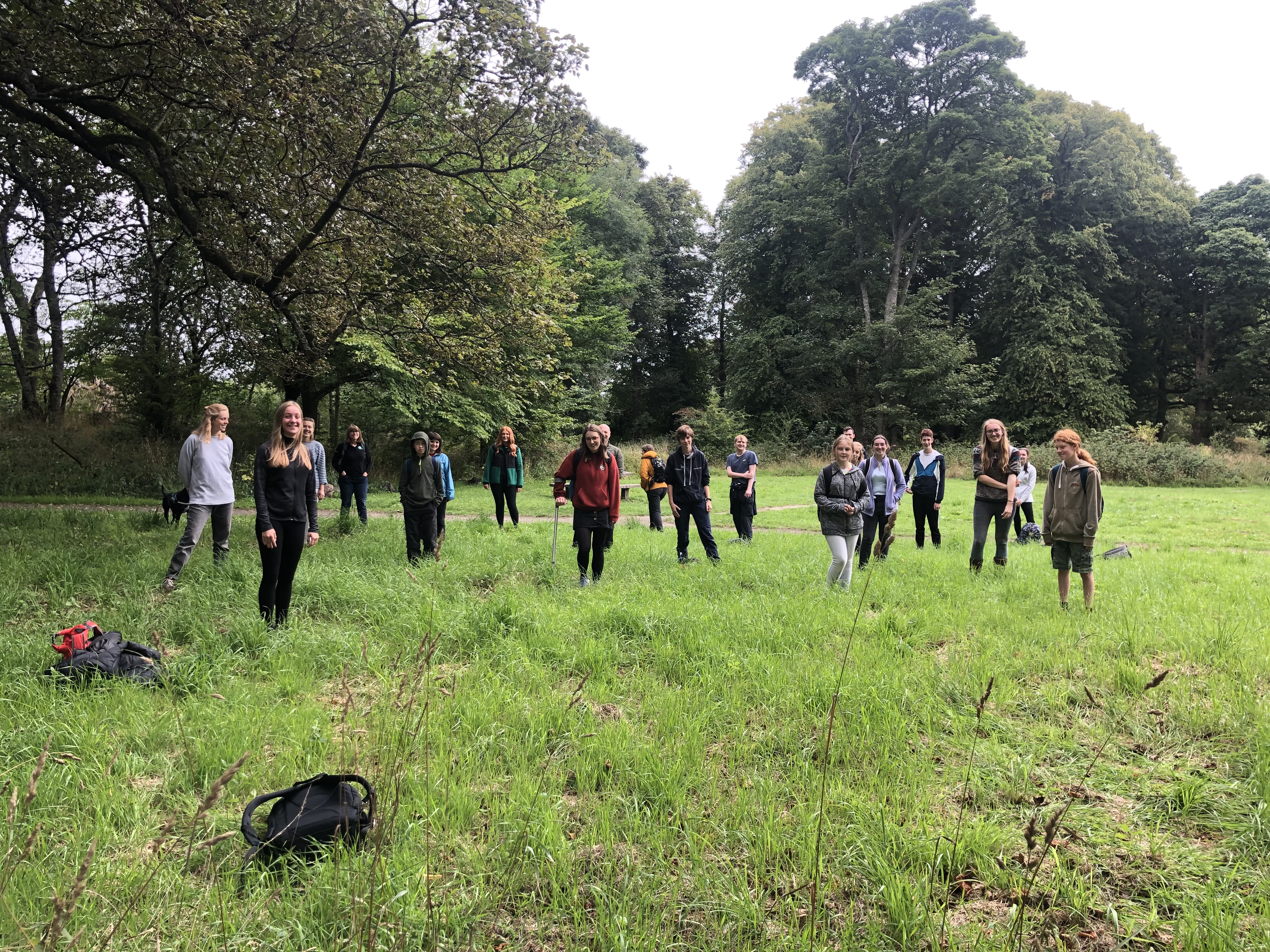 a group of young people gathered at spaced intervals in a green field surrounded by trees