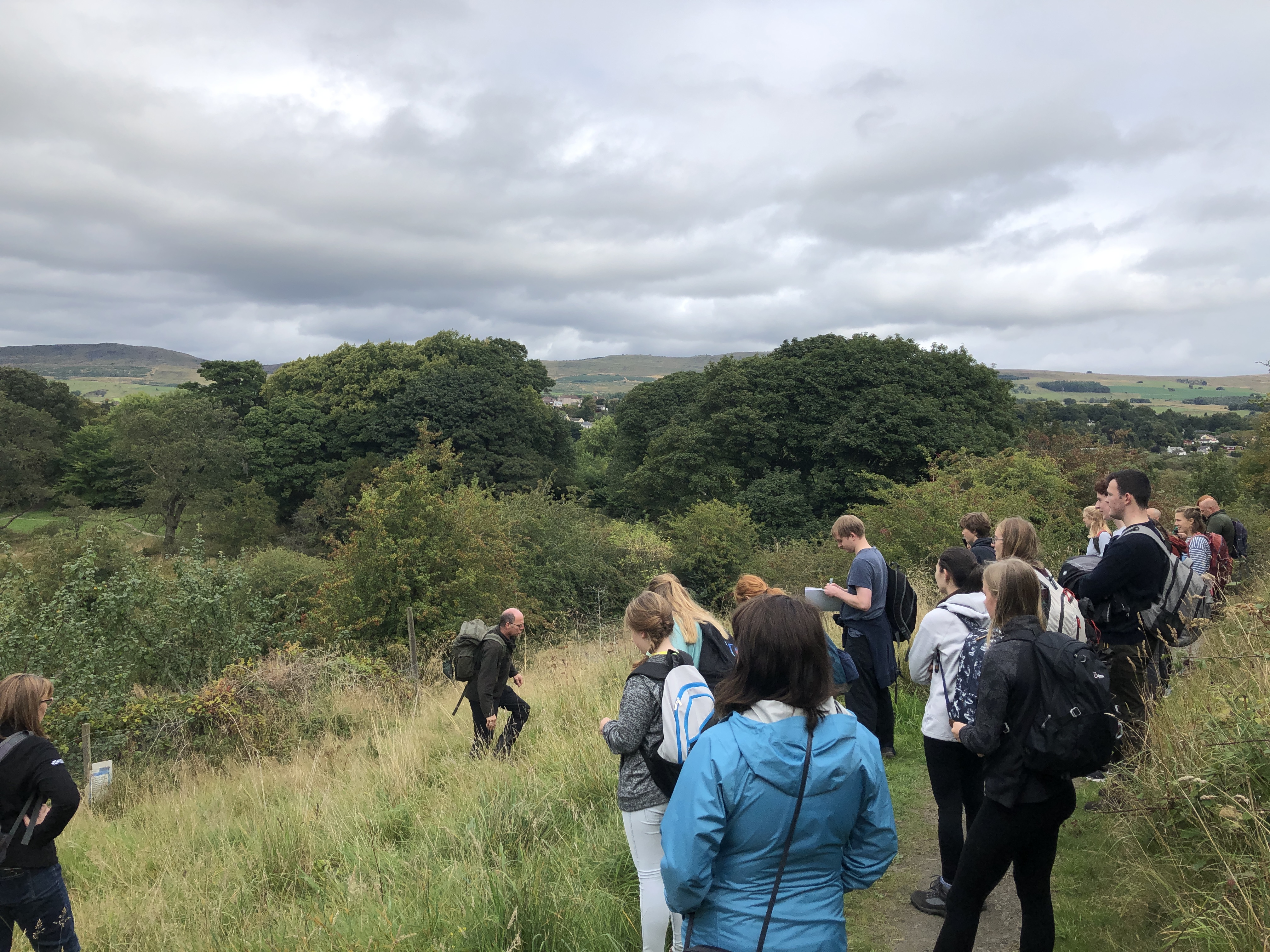 a group of young people standing on a slight grassy hill with a person in front of them who is teaching