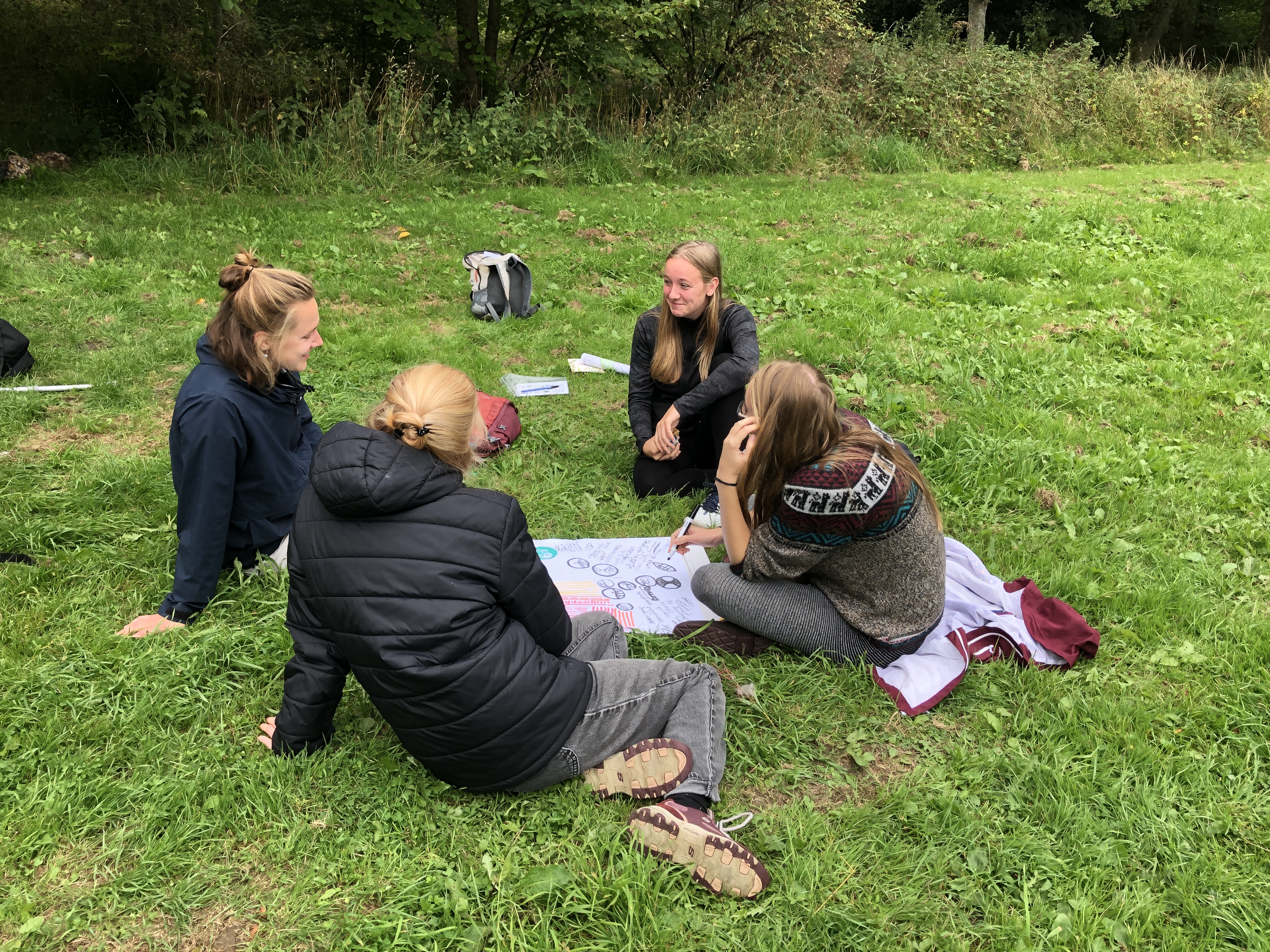 4 young people sit on the ground in a group with a piece of paper in front of them which has drawings and writing on