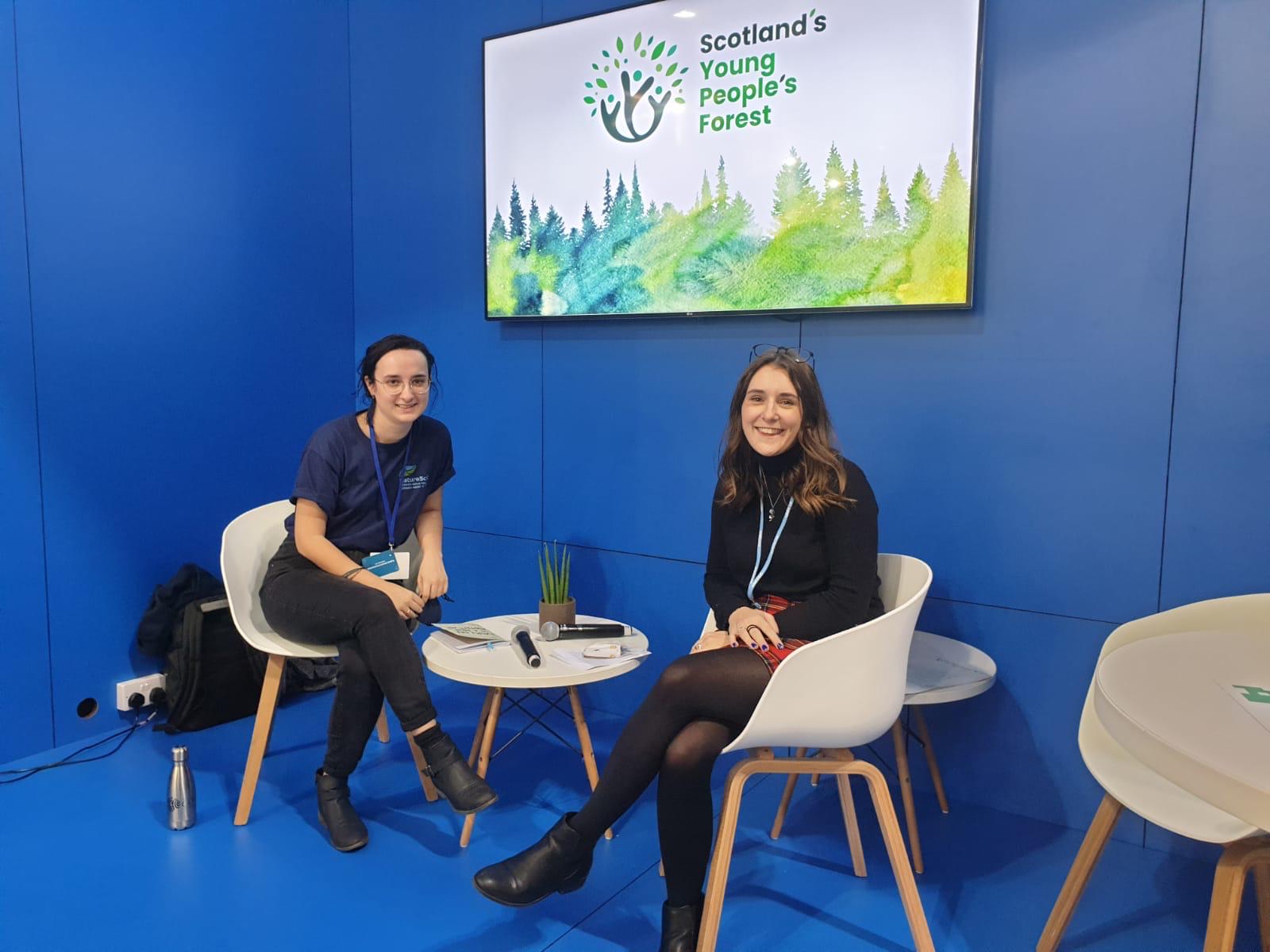 Emma and a member of NatureScot sit at a table with the SYPF logo on a screen behind them